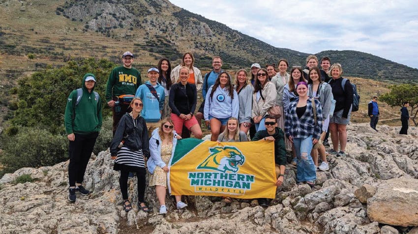 A group of students from NMU pose for a photo in Greece, during an IVHQ study abroad program; holding a flag with the text, Northern Michigan Wildcats