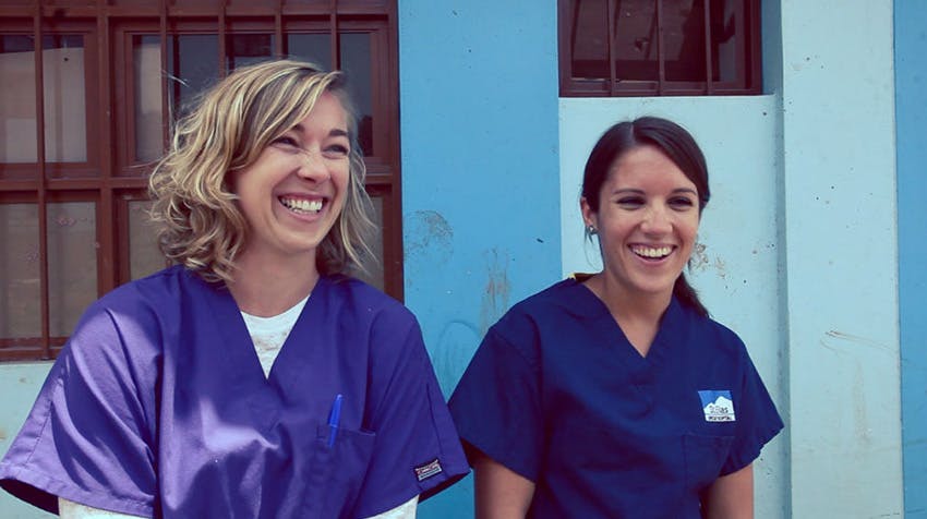 Two female volunteers in scrubs on a medical volunteer project smile and laugh together, standing against a blue and white wall