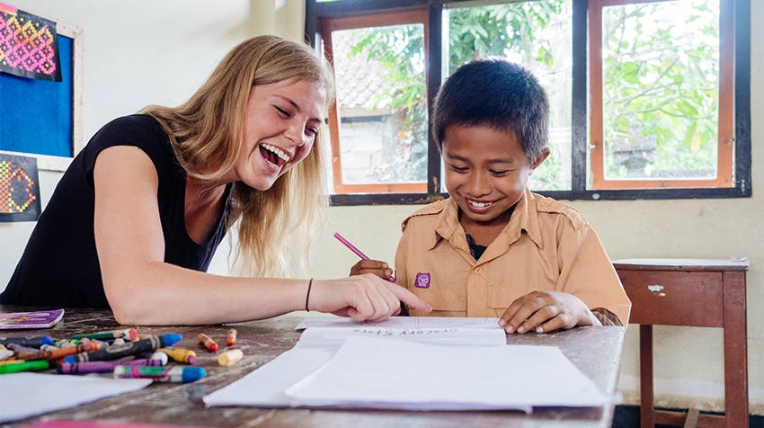 A teaching volunteer and young boy share a joyful moment in a classroom, engaged in drawing and learning together. Both are smiling with colorful art supplies