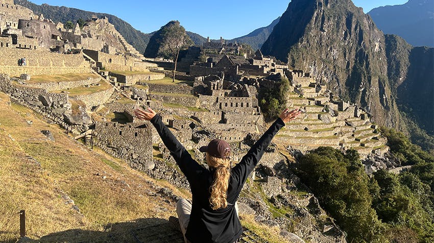 A volunteer traveller with long hair sits on a stone structure at Machu Picchu in Peru, arms raised in celebration. The ancient Incan city is visible in the background, surrounded by lush mountains and a clear blue sky