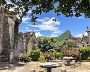 A charming garden courtyard with stone tables and benches, surrounded by flowering bushes and mountains in the background.