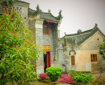 The exterior of a historic Chinese guesthouse with ornate tiled roofs, brick walls, and lush gardens in front.