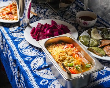 A buffet table with a variety of dishes, including noodles in a savory broth, vibrant cubes of dragon fruit, green leafy parcels, and sautéed vegetables.
