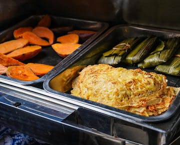 Assorted hot dishes in metal trays, including sliced baked sweet potatoes, banana leaf-wrapped parcels, and a stack of golden, pan-fried savory pancakes.