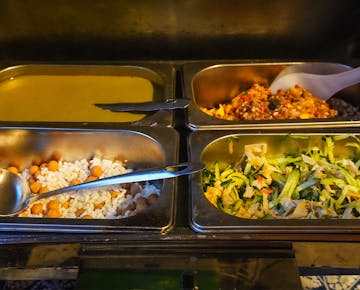 A selection of buffet-style dishes in metal trays, including creamy soup, stir-fried shredded cucumber and tofu, mixed beans and rice, and a spicy vegetable relish.