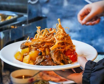 A plate of hearty food being served, topped with crispy fried vegetable strips, roasted potatoes, braised meat, and sautéed tomatoes.