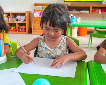 A young girl focuses intently while drawing at a green table during a classroom activity.