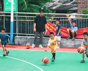 Young boys enthusiastically dribble basketballs on a village court, while a volunteer supervises the activity.