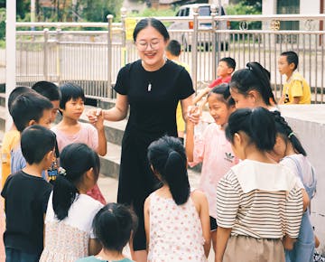 A smiling teacher holds hands with children in a circle, leading an outdoor group activity.