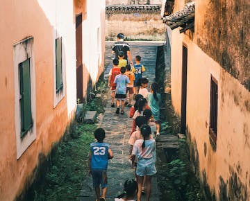 A group of children walks in a line down a village alleyway, following their teacher to the next activity.