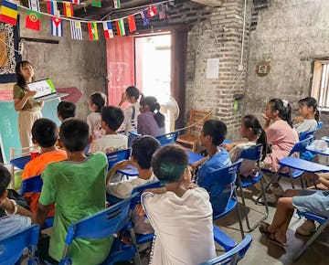 A volunteer stands at the front of a rustic classroom, holding a whiteboard and engaging children seated on blue chairs under international flags.