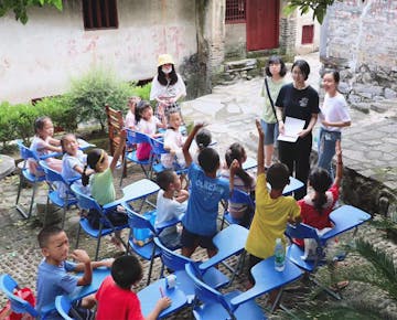 Children seated on blue chairs in an outdoor courtyard classroom, eagerly raising their hands as volunteers lead the lesson.