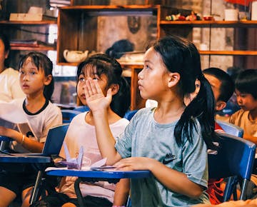 A student raises her hand while attentively listening in a classroom filled with engaged children.