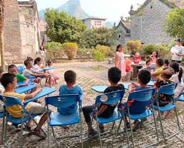 Children and volunteers gather outdoors in a scenic courtyard, with a mountain view in the background, participating in a lesson.
