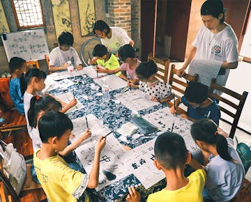 Children sit around a long table, practicing Chinese calligraphy with brushes under the guidance of volunteers.