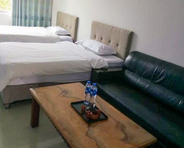 A bright bedroom with two beds, a black leather sofa, and a wooden coffee table with bottled water.