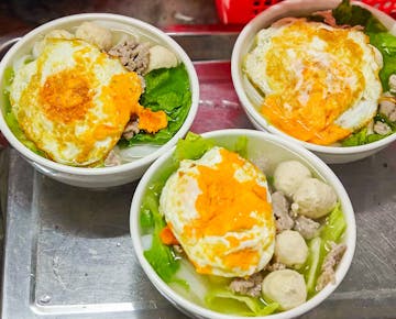 Three bowls of noodle soup topped with fried eggs, leafy greens, meatballs, and sliced pork, served on a metal tray.