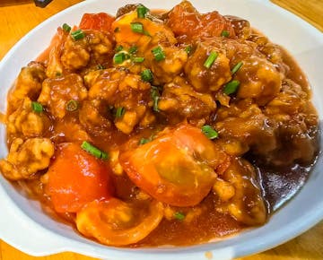 A serving dish of sweet and sour pork with tomatoes and green onions in a rich, glossy sauce.