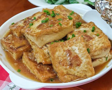 A bowl of golden-brown tofu squares in a savory brown sauce, garnished with chopped green onions.
