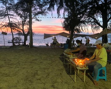 People grill food at outdoor barbecues on a sandy beach at sunset, with tents, string lights, and the sea in the background.