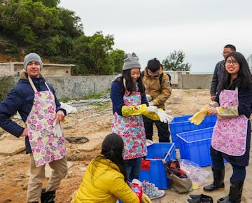 A group of smiling volunteers wearing aprons, rubber gloves, and winter hats prepare for a conservation task next to blue bins outdoors.