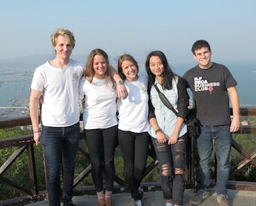 Five young volunteers stand arm-in-arm, smiling on a hilltop with the sea and coastline in the distance.