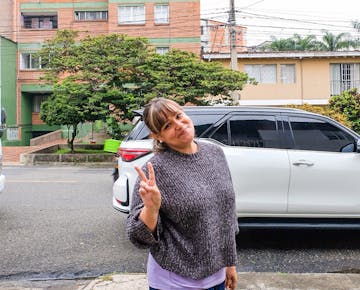 A woman standing on a residential street making a peace sign and smiling at the camera. She is wearing a grey sweater and standing in front of a white SUV, with houses and greenery in the background.