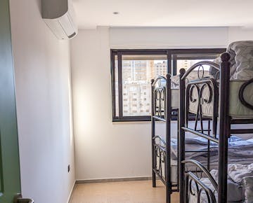 A bright volunteer room featuring bunk beds, tiled floors, and a large window with city views, complemented by air conditioning on the wall.