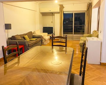 A shared living room with sofas, a dining table, a small TV, and a sliding glass door opening to a balcony, creating a relaxed community space.