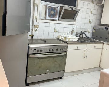 A compact kitchen with tiled walls, a gas oven, refrigerator, stainless steel sink, and dish rack, set up for volunteer self-catering.