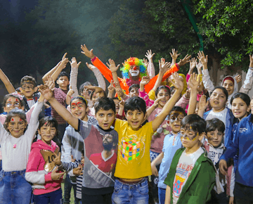 A large group of excited children with face paint and costumes smile and cheer outdoors during a festive evening celebration.