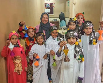 A group of children dressed in colorful traditional clothing proudly display handmade decorations alongside a smiling local teacher.