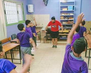 A volunteer leads a group of boys through an active movement session in a classroom, with raised arms and big energy all around.
