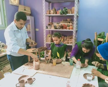 A teacher and several children engage in a pottery-making session, shaping clay items in a cheerful classroom filled with handmade ceramics.
