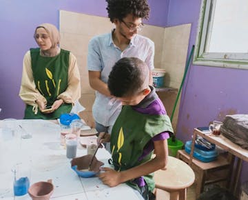 A volunteer helps a child shape a clay bowl during a hands-on art session in a purple-walled classroom, with paints and tools spread out on the table.