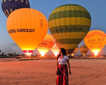 A woman stands on a desert plain at dawn, admiring vibrant hot air balloons preparing for take-off near Luxor, Egypt.