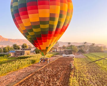 A hot air balloon prepares for lift-off over lush farmland in Luxor at sunrise, with golden light illuminating the landscape.