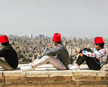 Three young men wearing red fezzes sit on a stone wall, gazing out over the cityscape of Cairo on a clear day.
