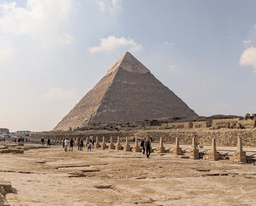 Tourists walk toward the iconic Pyramid of Khafre at the Giza Plateau, surrounded by ancient stone ruins and desert views.