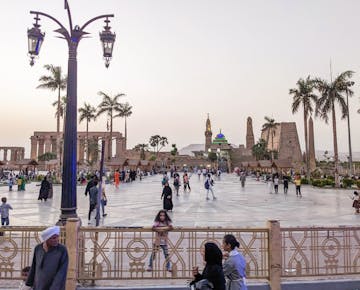 Locals and tourists gather in a lively square outside the illuminated Luxor Temple during twilight, with palm trees and minarets in the background.