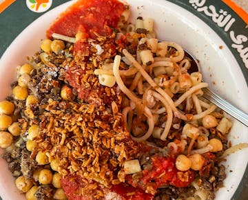 A colorful bowl of Koshari featuring pasta, lentils, chickpeas, fried onions, and tomato sauce.