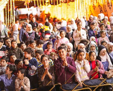 A large group of smiling children sit and stand in front of a lit-up festival stage, clapping and watching a live performance under glowing lights.