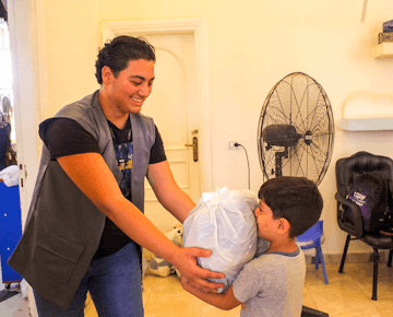 A smiling young man hands a large plastic-wrapped donation bundle to a child inside a brightly lit room, fostering community support and connection.