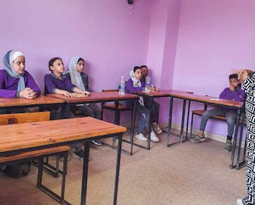 A group of primary-aged students attentively listen to a female teacher in a purple-walled classroom, seated at wooden desks.