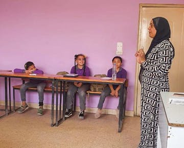 A woman stands at the front of a cheerful classroom, engaging students in discussion while children respond with interest and smiles.