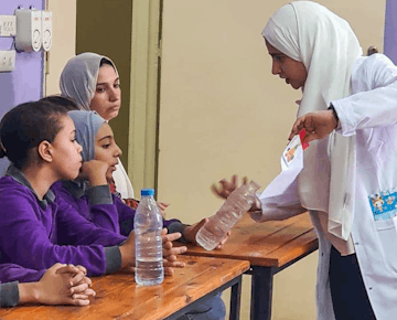 A teacher in a white coat holds a flashcard and bottle while explaining a lesson to young girls seated at desks, enhancing their understanding through demonstration.