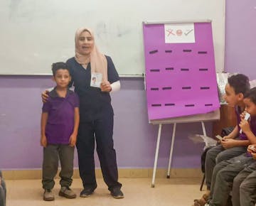A smiling teacher stands beside a young student at the front of the classroom, holding a flashcard while classmates watch during an English practice activity.