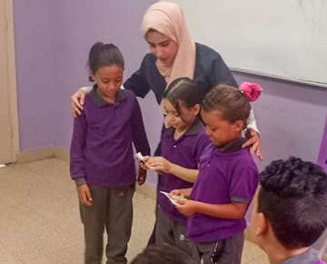 A female volunteer encourages a small group of girls as they participate in a classroom reading activity using word cards.