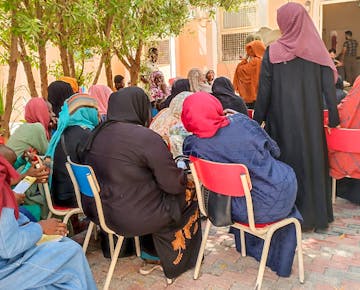 A group of women in colorful hijabs gather outdoors for a community session, seated under trees in a shaded courtyard.
