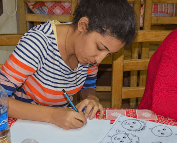 A young woman sketches cartoon faces during a creative class session, concentrating intently on her pencil work at a wooden table.